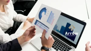 Two professionals reviewing charts and graphs, with one holding a printed report and a laptop displaying a bar chart with Arrow Dispatch Services LLC logo