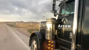 Close-up view of a black semi-truck with the Arrow Dispatch Services LLC logo on the door, parked on a rural highway with open plains and cloudy sky in the background.