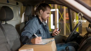 Delivery driver sitting in a van checking his smartphone while filling out paperwork on a clipboard, with a cardboard box on his lap and the Arrow Dispatch Services LLC logo visible on the right.