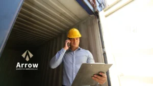 Freight broker from Arrow Dispatch Services checking shipment details on a clipboard while talking on phone at a shipping container.